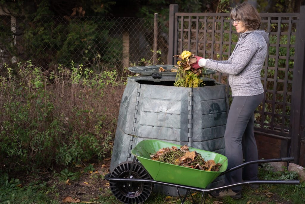 Woman adding garden waste into a plastic compost bin with a wheelbarrow nearby.