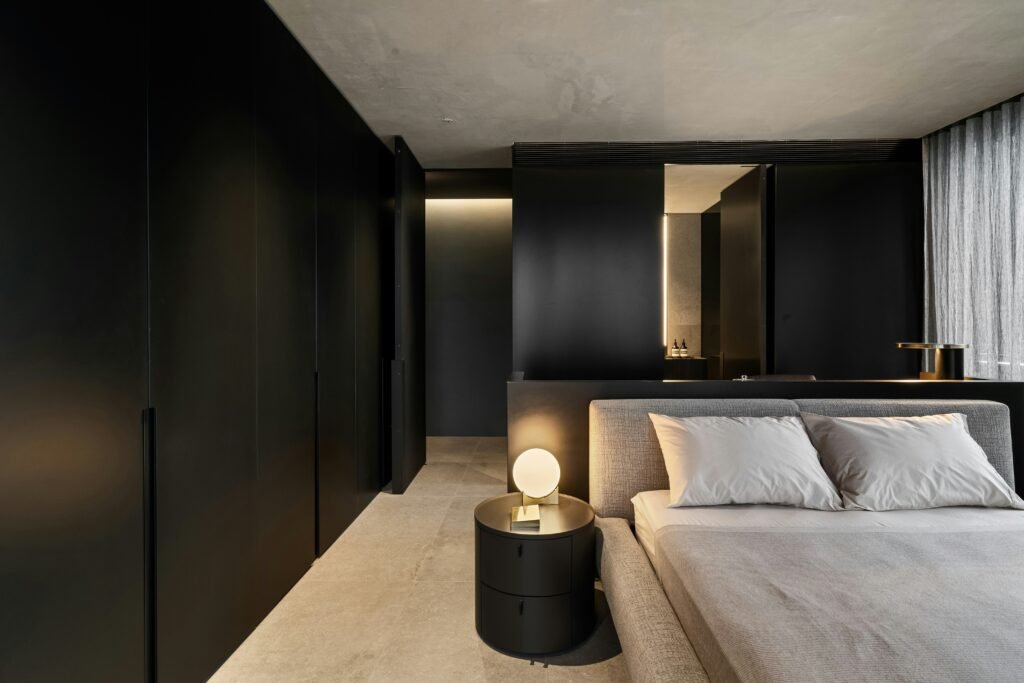 Minimalist modern bedroom with black wall panels, gray upholstered bed, and warm ambient lighting.